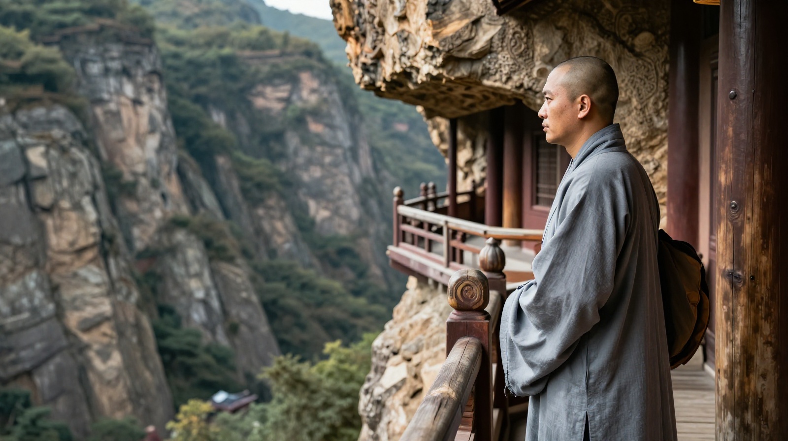 A monk in traditional grey robes standing on the wooden walkway of the Hanging Temple in Shanxi, China, overlooking a mountain valley.