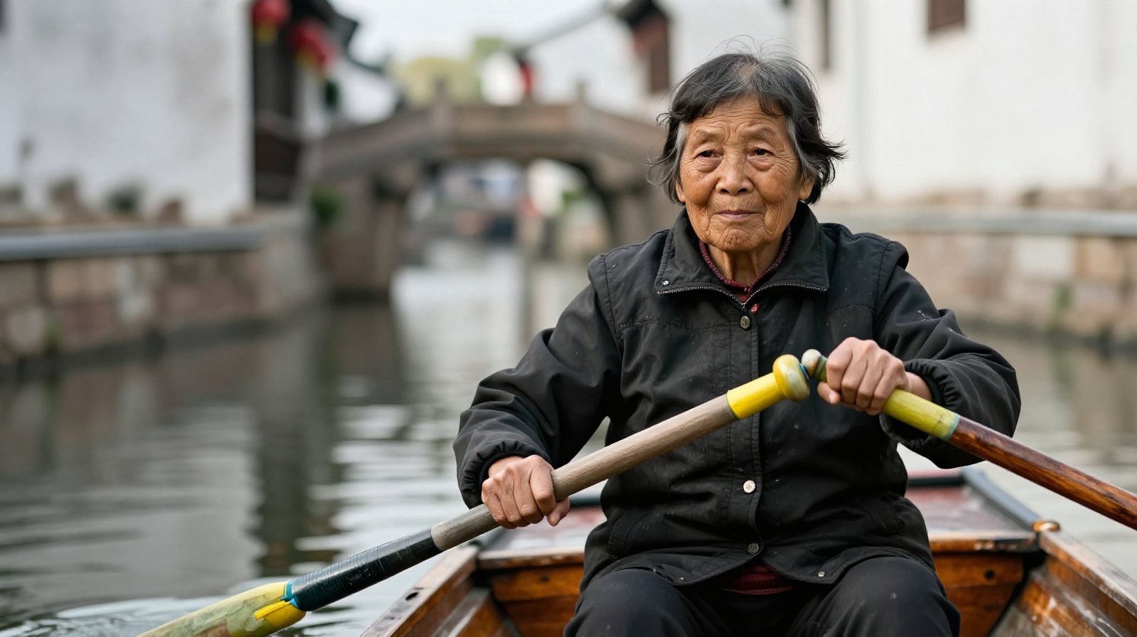 Elderly Chinese woman rowing a traditional black-hulled boat through a quiet canal in Suzhou water town at sunrise
