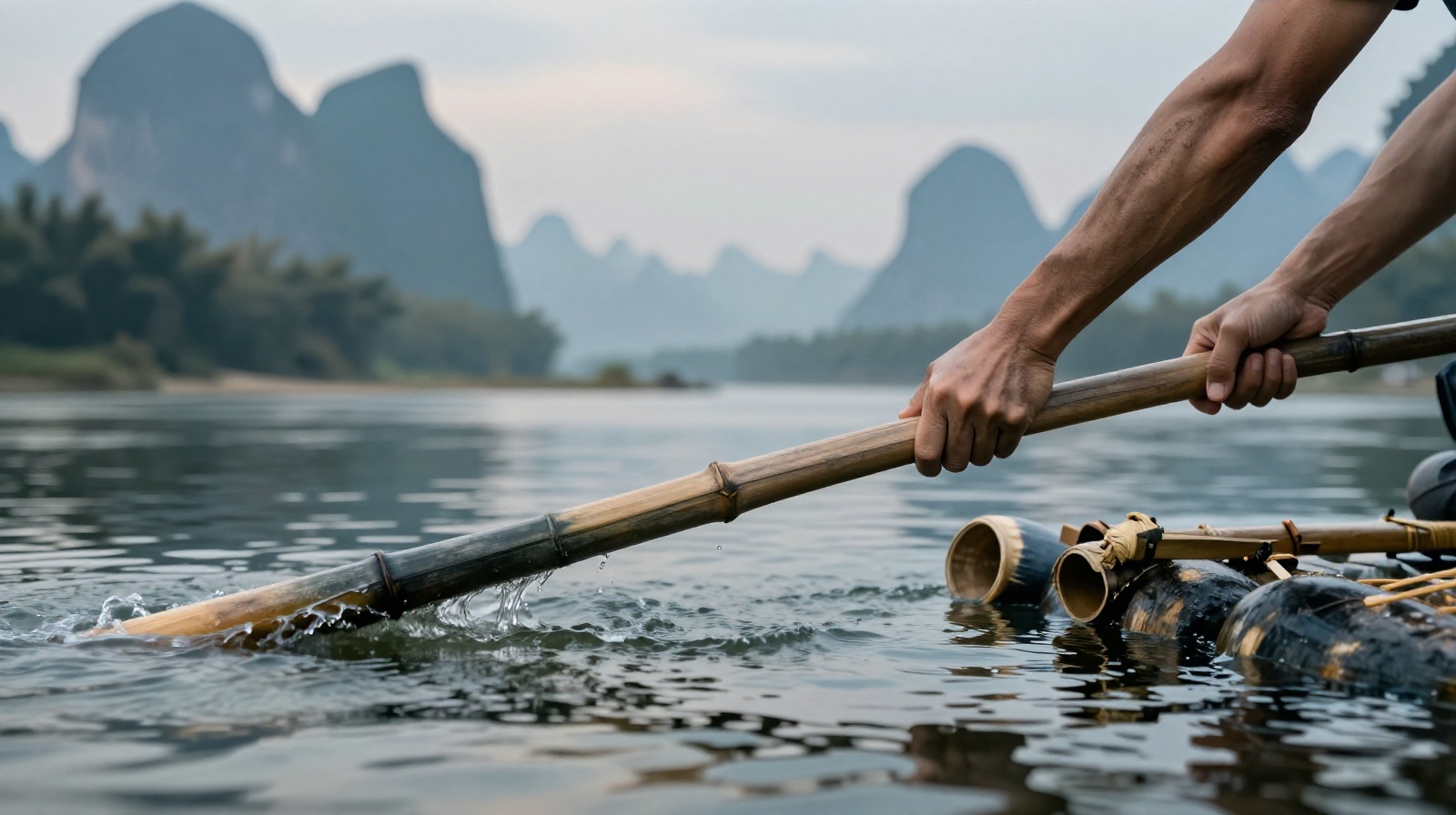 A close-up view of a local boatman's weathered hands gripping a long wooden bamboo pole while navigating a raft on the Yulong River during sunrise, with misty karst peaks in the background