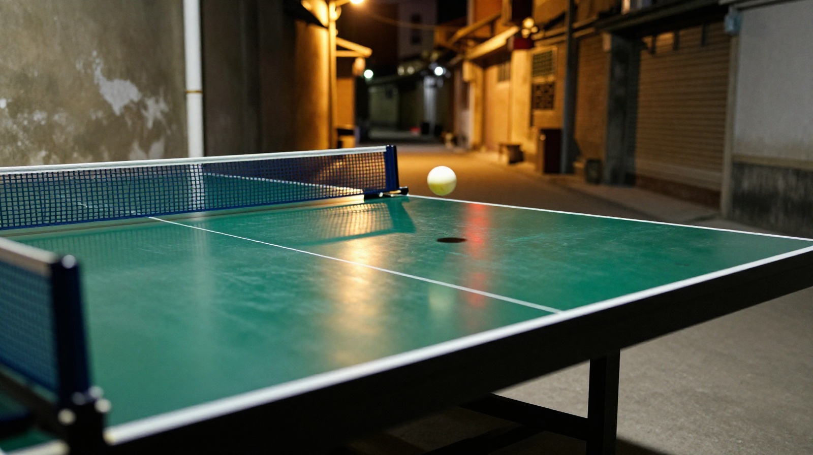 A close-up view of a green table tennis table in a dimly lit Chinese neighborhood park at night with a ball in mid-air capturing the action of a game