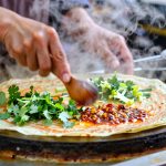 A Chinese street food vendor preparing a savory jianbing crepe for customers early in the morning with visible steam rising from the cooking station