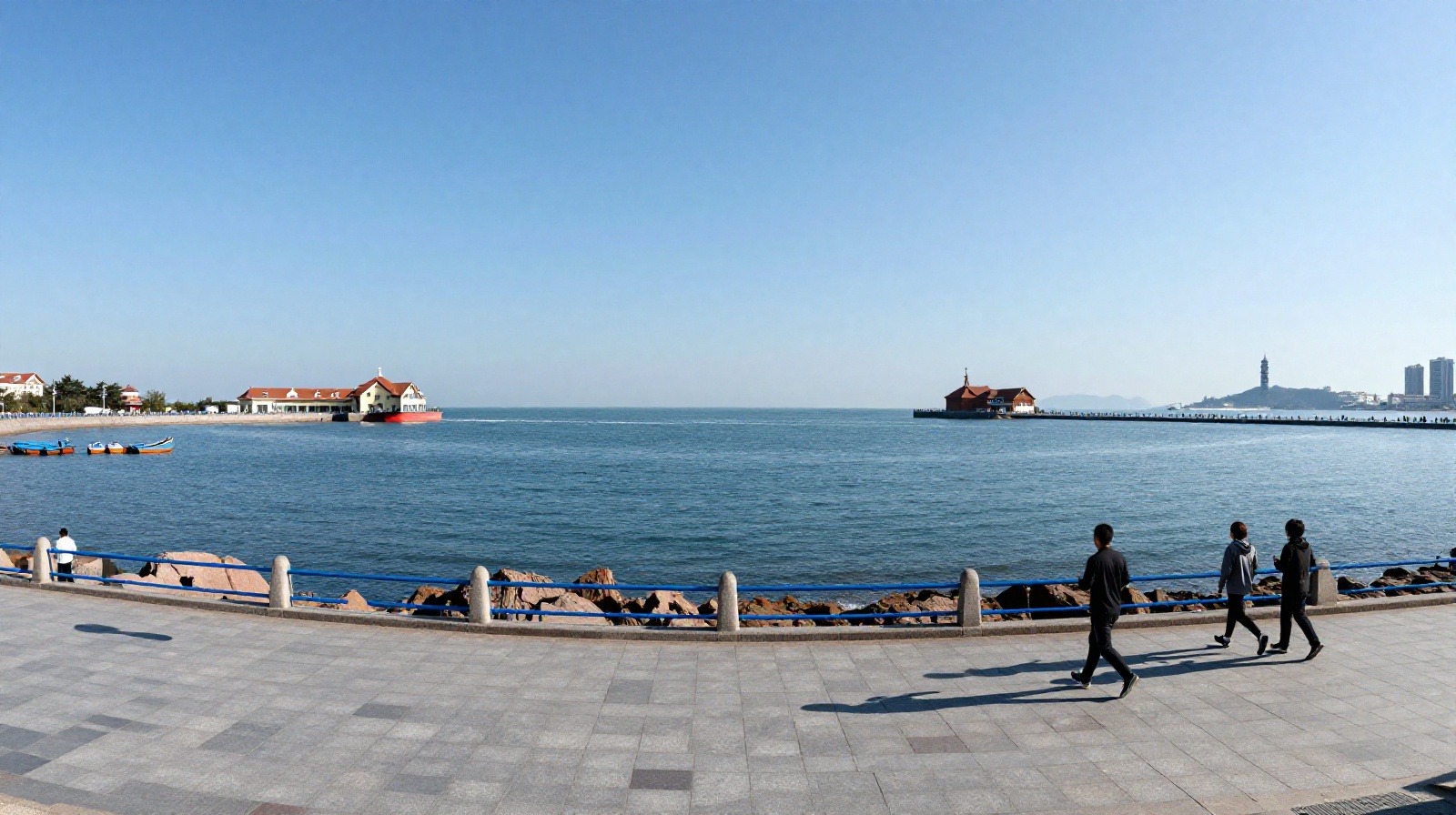 People enjoying a walk along the coastal promenade in Qingdao with the historic Zhan Qiao Pier in the background