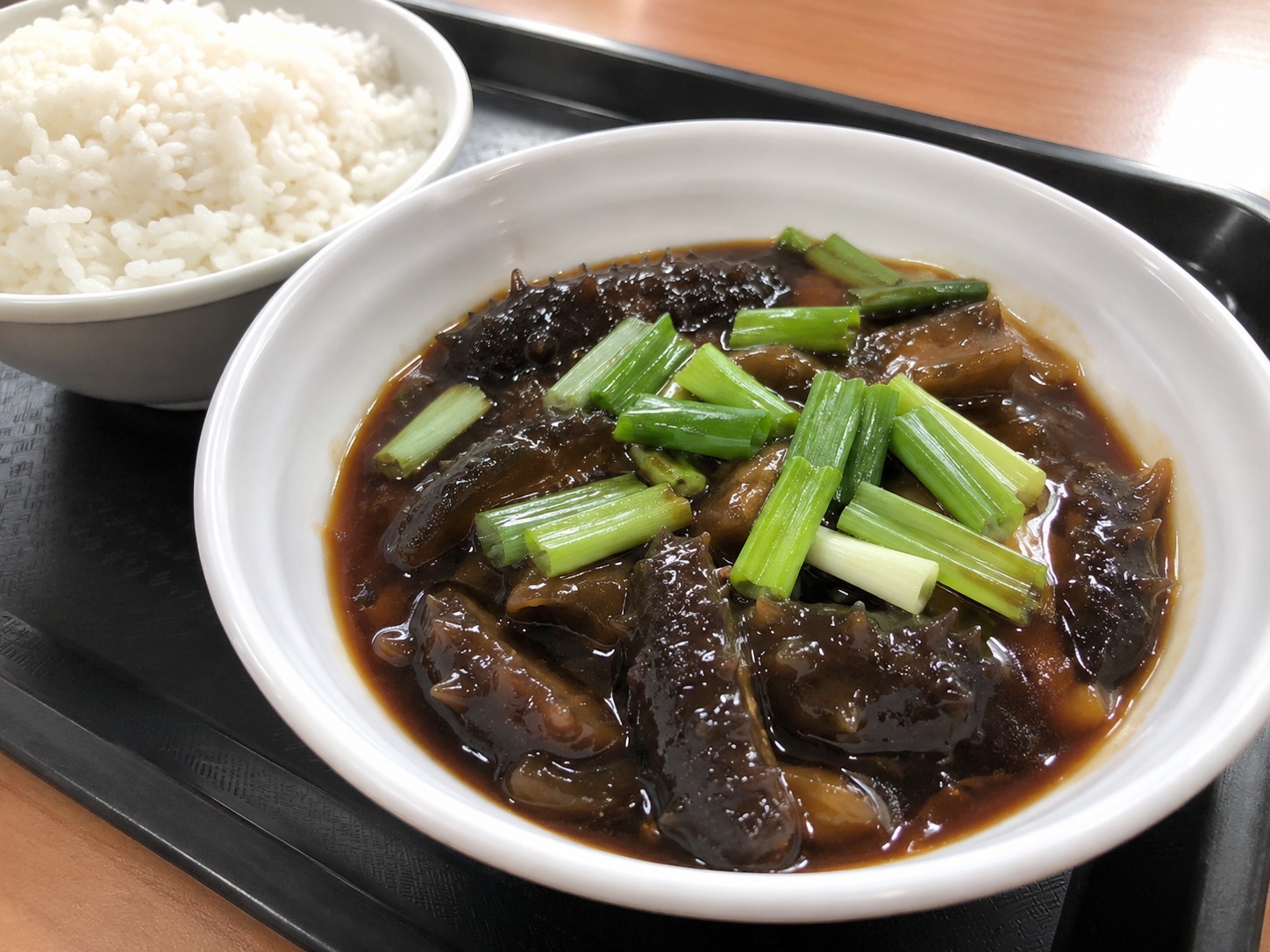 A close-up photo of Braised Sea Cucumber with Scallions and white rice served in a standard Chinese community canteen meal tray.