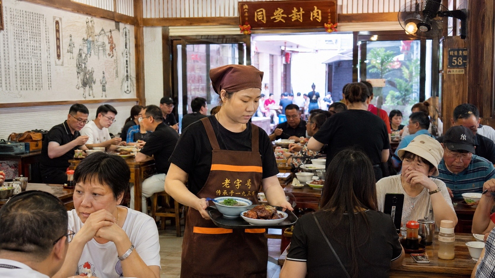 Diners at a Xiamen restaurant sharing tables, with servers carrying traditional Fujian dishes like seafood soup and braised pork during the midday rush.