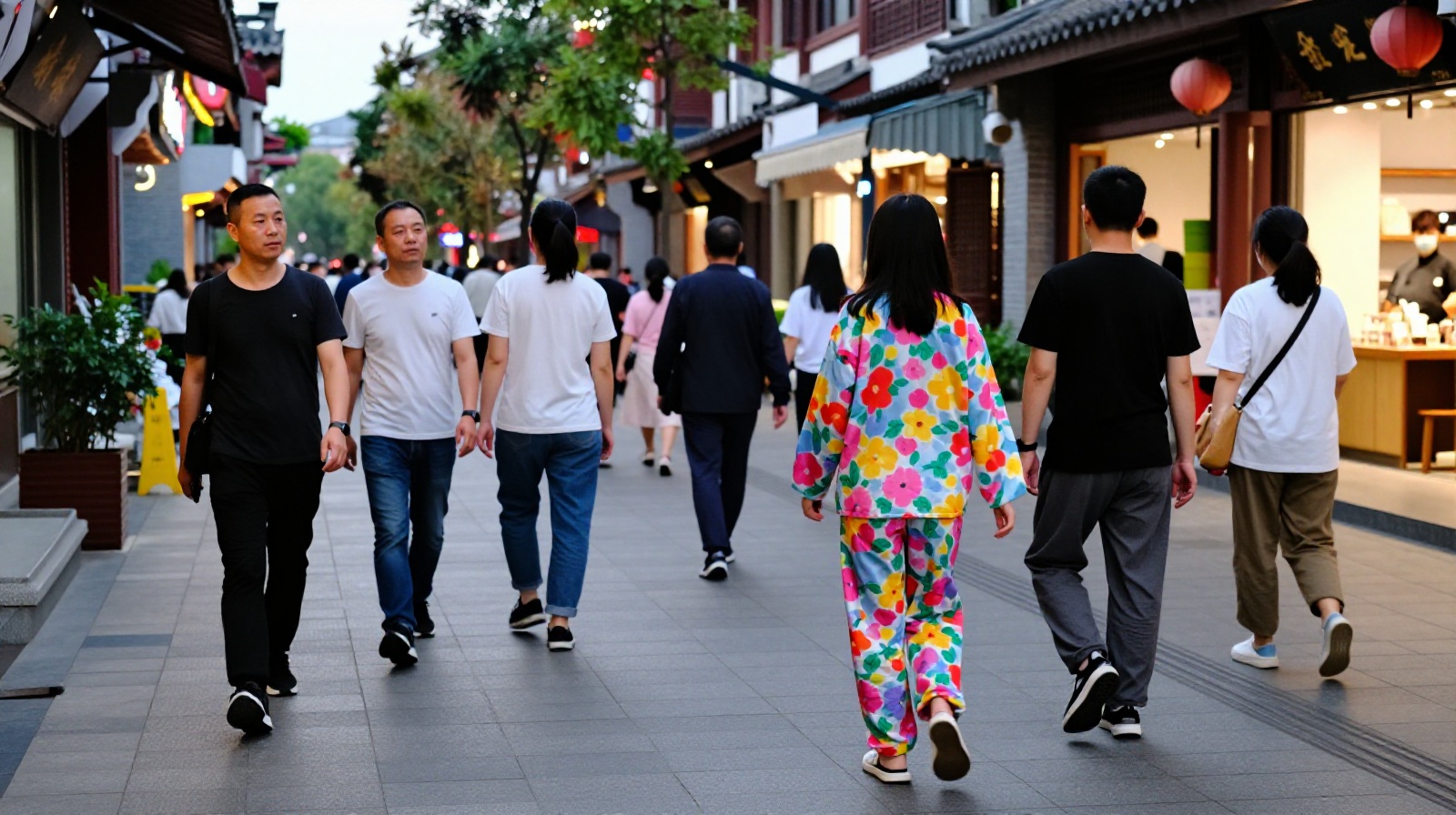 Pedestrians on a street in southern China, including a person wearing colorful floral pajamas, walking past local shops.