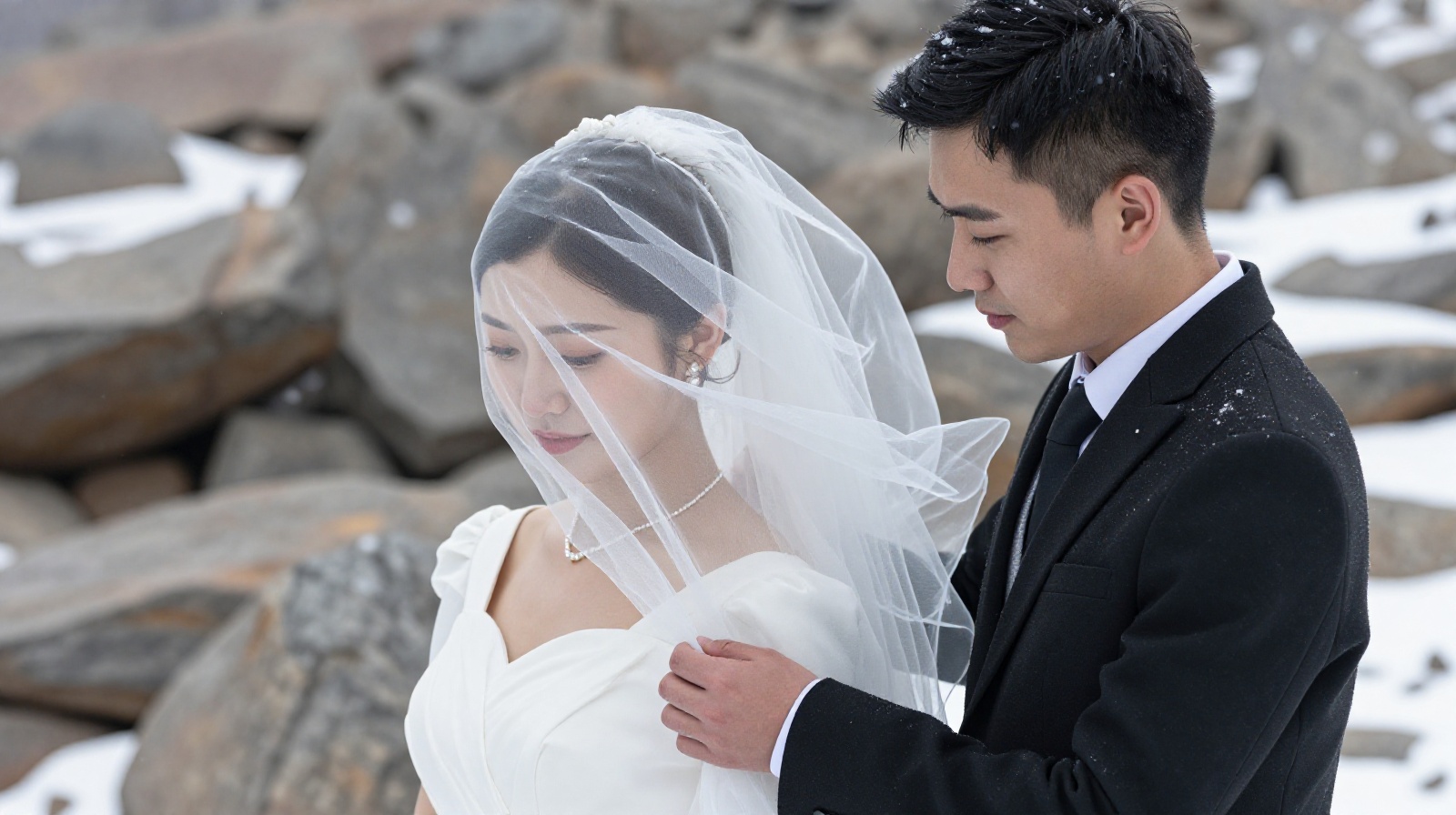 A close-up of a Chinese bride and groom in wedding clothes standing on a snowy mountain, with snowflakes falling and wind blowing their attire.