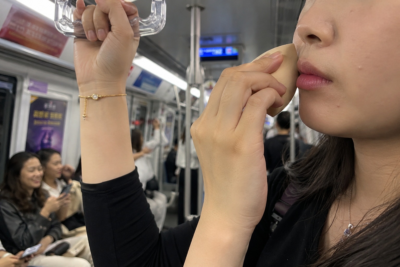 A close-up view of a commuter applying makeup on a moving subway train, using an overhead handle for balance.