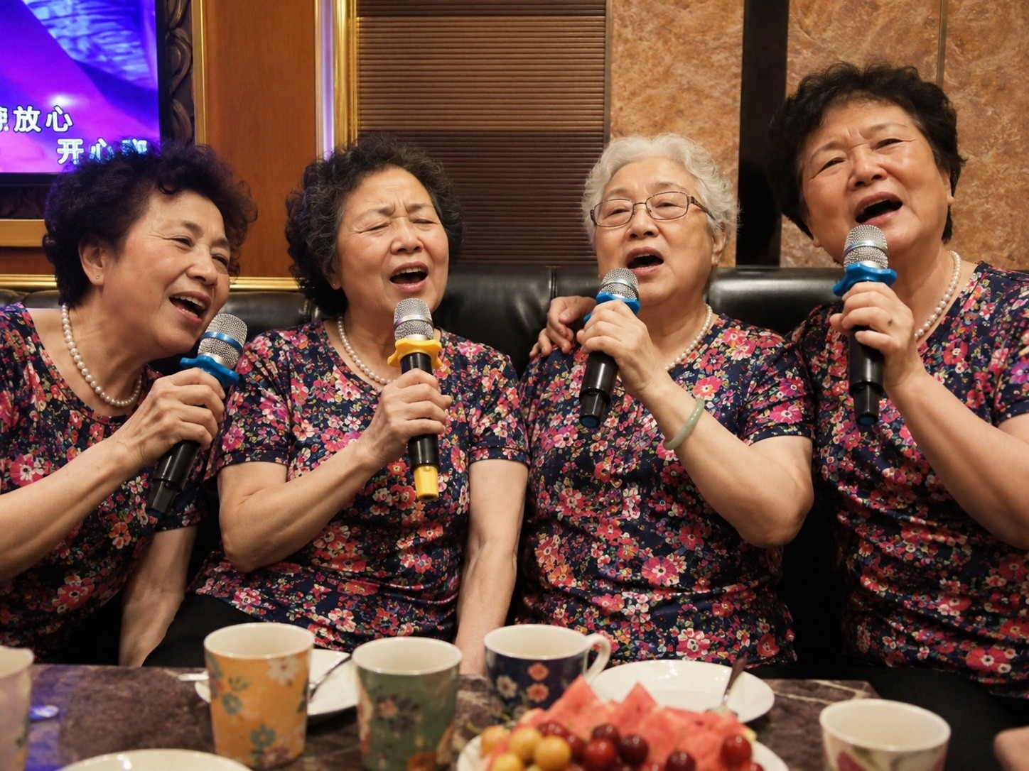 Elderly Chinese women singing karaoke in a private room during the daytime, wearing floral shirts and enjoying tea.