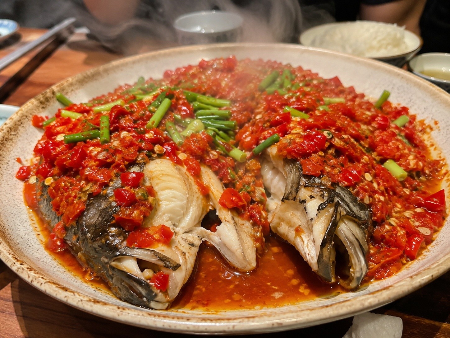 Close-up of a steaming plate of Chopped Chili Fish Head covered in red fermented chilies, being served at a Chinese restaurant.