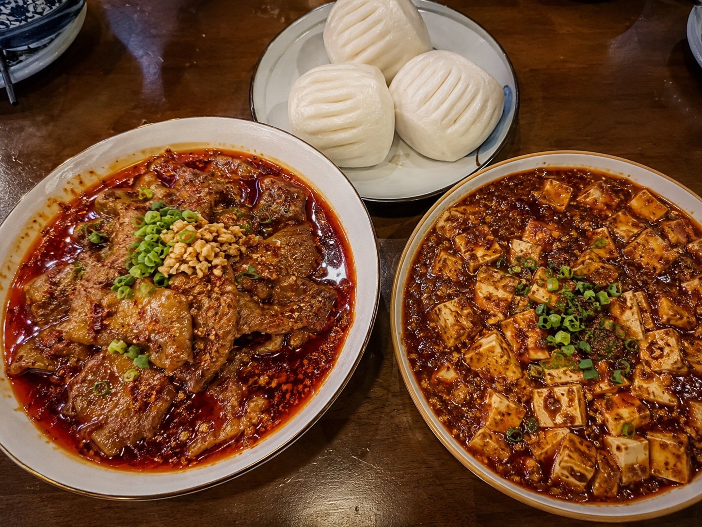 A table setting with classic Sichuan dishes including spicy beef slices, mapo tofu, and steamed buns in simple ceramic bowls.