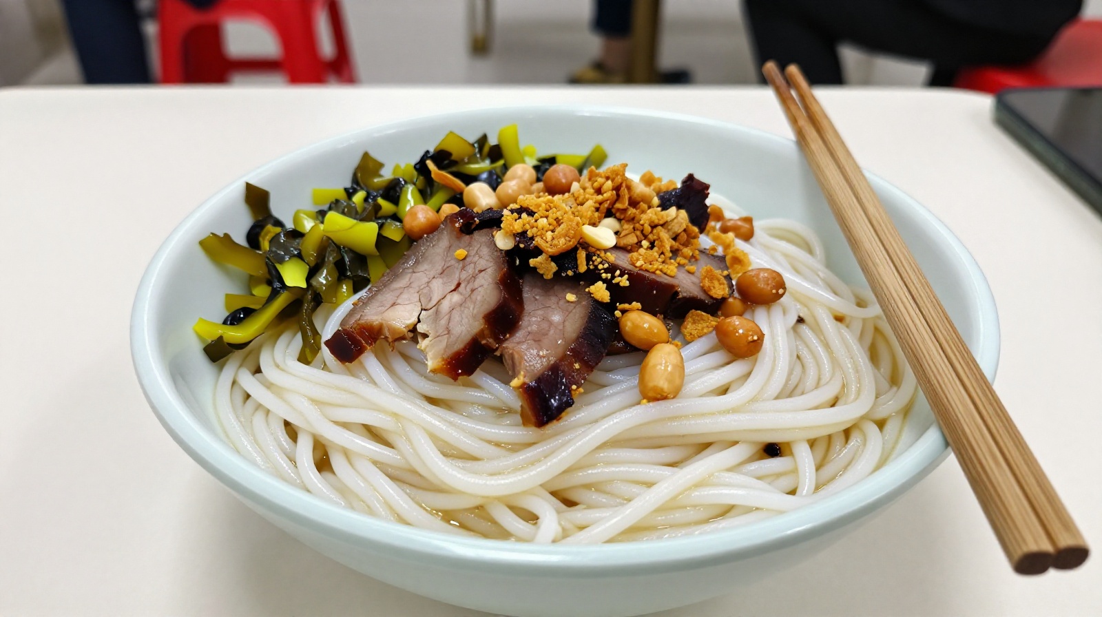 A close-up view of a bowl of Guilin rice noodles with toppings like peanuts, meat, and pickled vegetables on a plastic table
