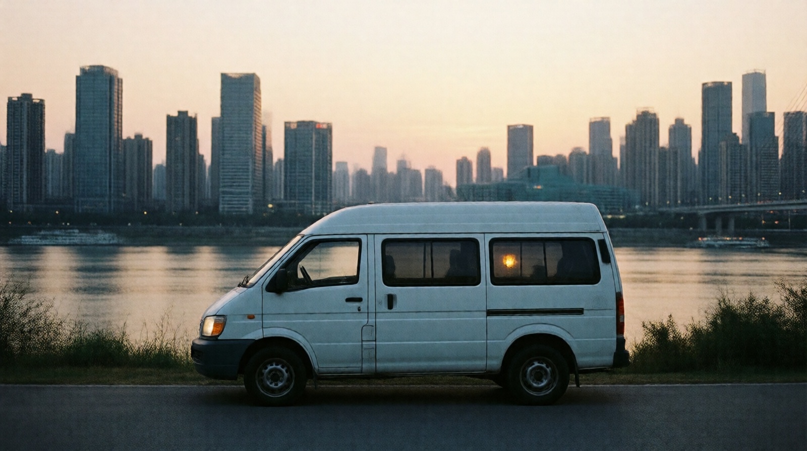 A white van parked by a river at dusk with a city skyline in the background, symbolizing the mobile lifestyle of Chinese construction workers.