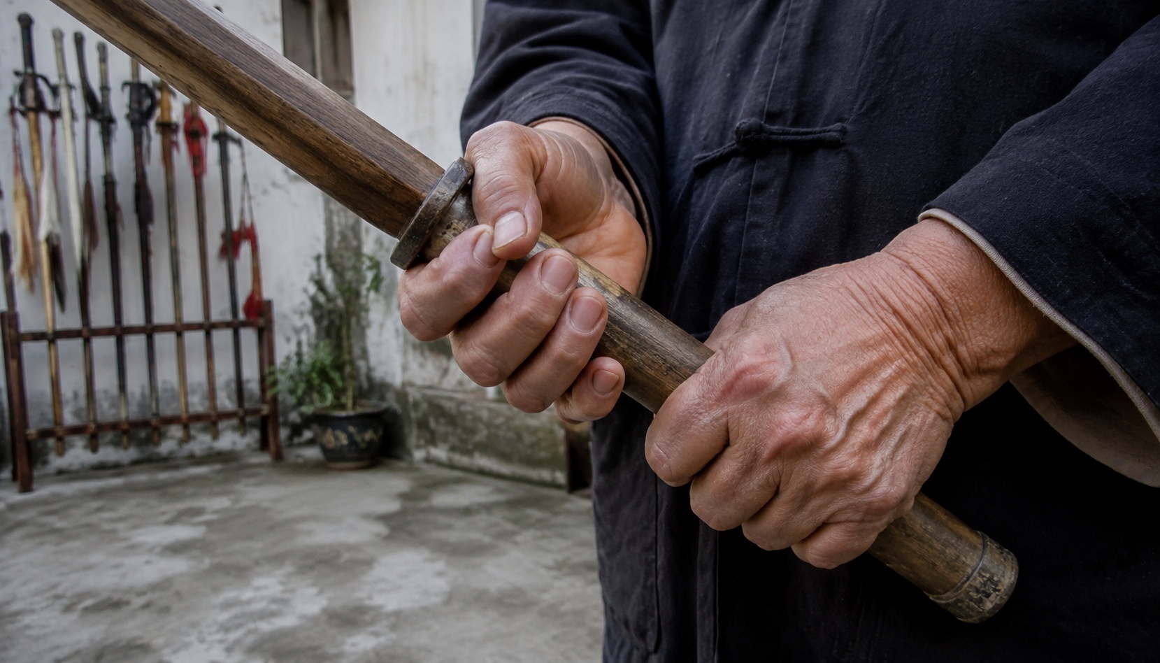 Elderly Chinese retired teacher's hands holding a wooden practice sword with calloused fingers, backyard martial arts equipment visible