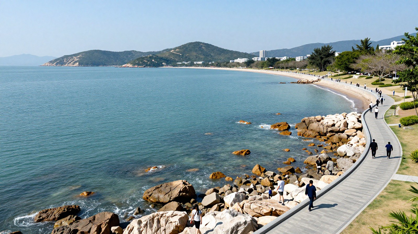 Scenic view of the Dapeng Peninsula coastline in Shenzhen, showing tourists walking along a coastal park trail with clear blue water and rocky shores.