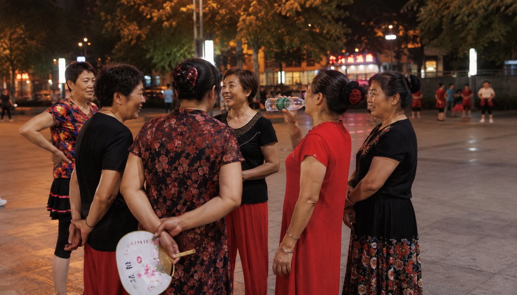 Chinese aunties socializing during a square dance break, laughing and talking in a public plaza at night