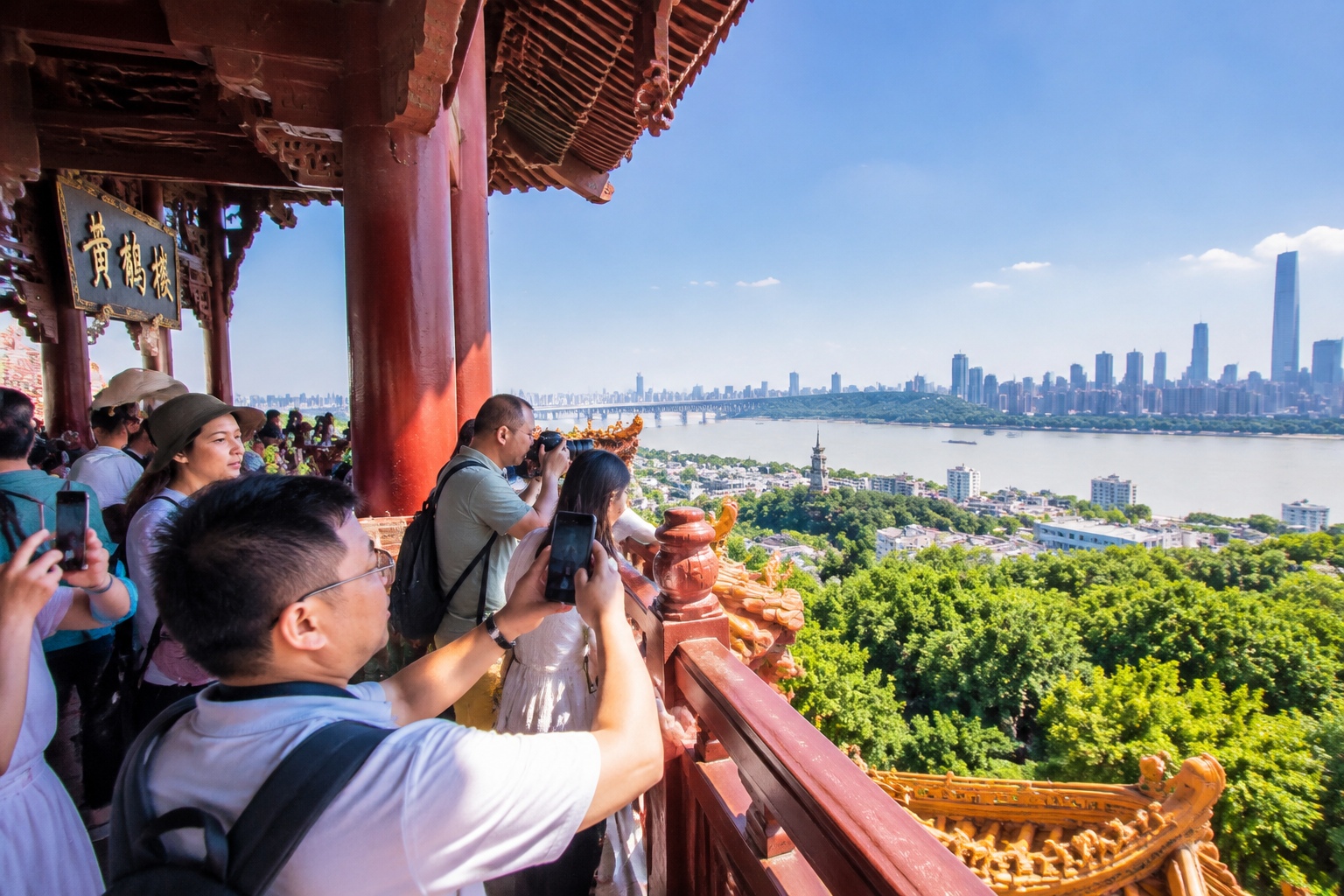 Tourists at the top of Yellow Crane Tower overlook the Yangtze River and Wuhan skyline, capturing the moment with phones.
