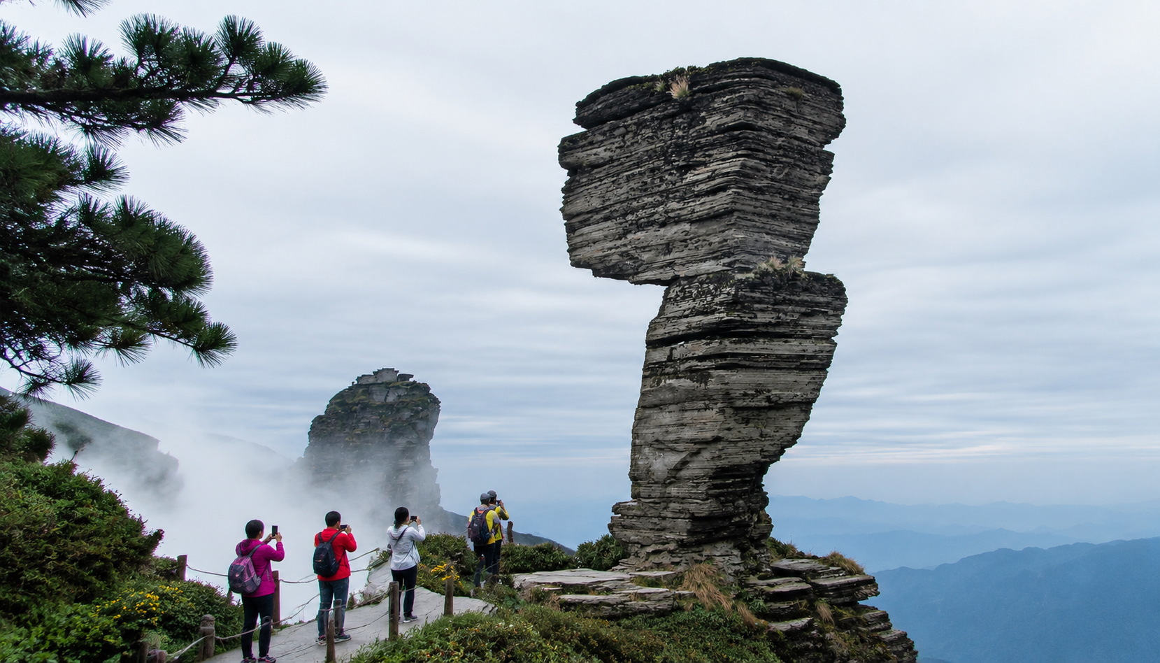 Hikers taking photos near Mushroom Rock, a famous stacked boulder on Fanjingshan hiking trail, with cloudy forest backdrop.