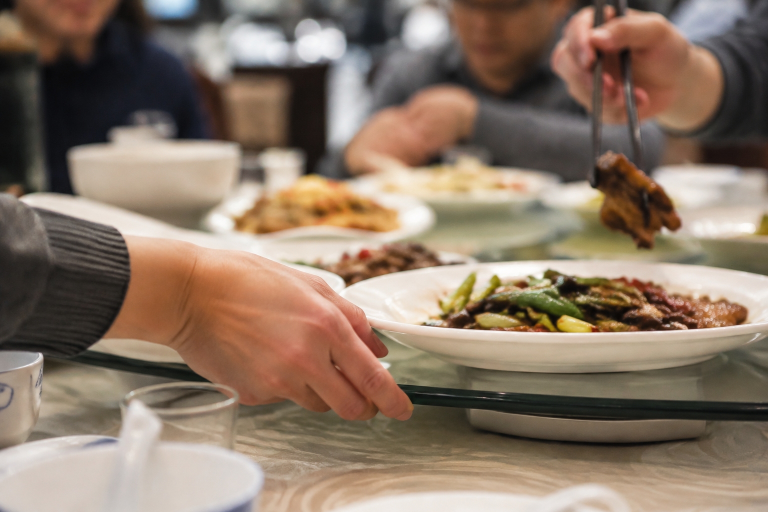 A diner carefully turns the rotating table while another guest uses chopsticks to pick up a dish, showing shared meal etiquette in China.