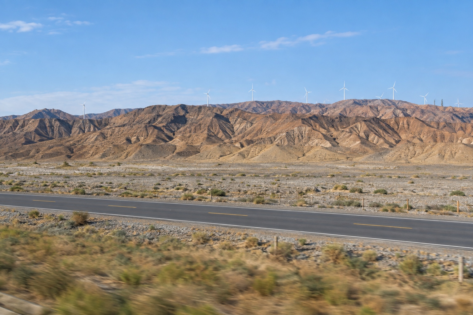 View from a moving train across the Hexi Corridor in Gansu with bare brown mountains, wind turbines and an empty desert road