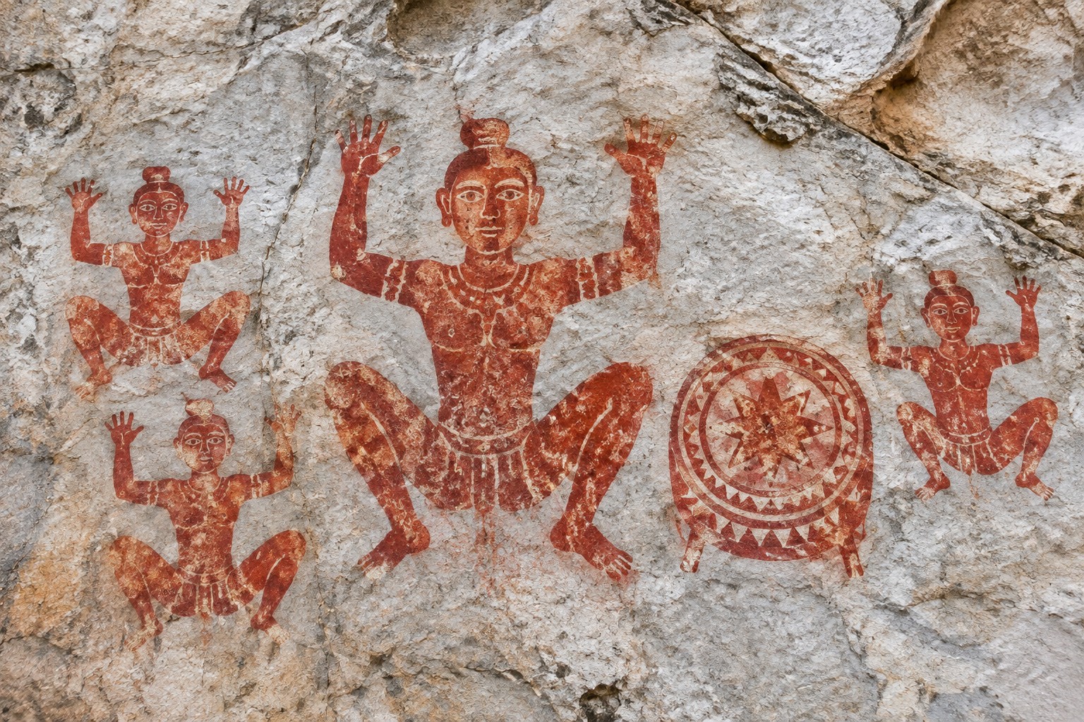 Close-up of ancient red ochre rock paintings on a limestone cliff, showing squatting human figures with raised arms beside a painted bronze drum with a star pattern